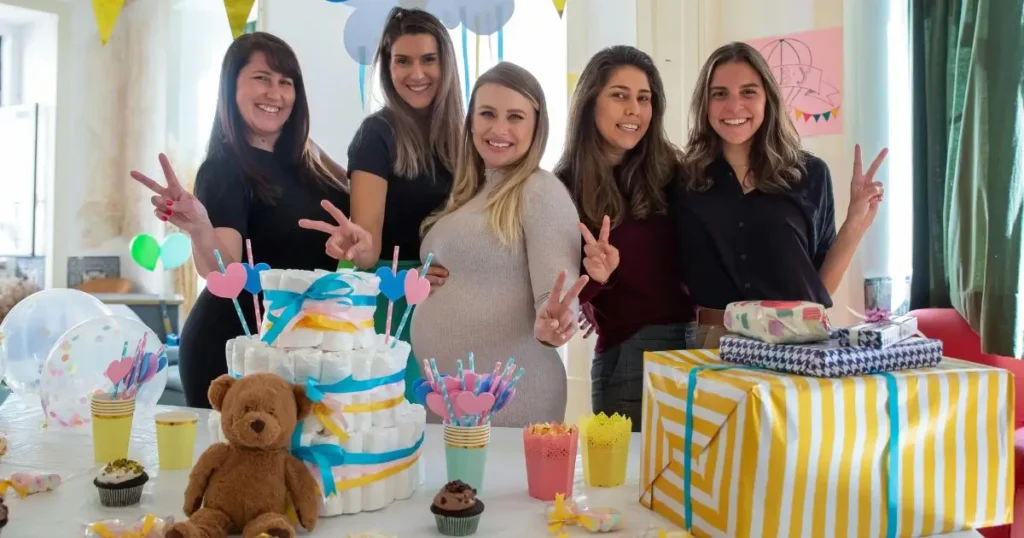 A group of five women posing at a baby shower celebration, with the mom-to-be in the center wearing a beige maternity dress. The table is decorated with a diaper cake, stuffed teddy bear, colorful cups, and wrapped gifts. The atmosphere is cheerful with balloons and banners in the background